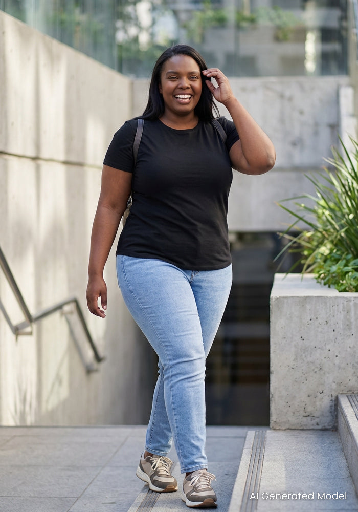 A woman with dark hair smiles as she walks outdoors on a ramp, wearing the Woolx Liza Crew Neck Tee in black, light blue jeans, and sneakers. She holds her hair and carries a backpack; plants and concrete walls are in the background.