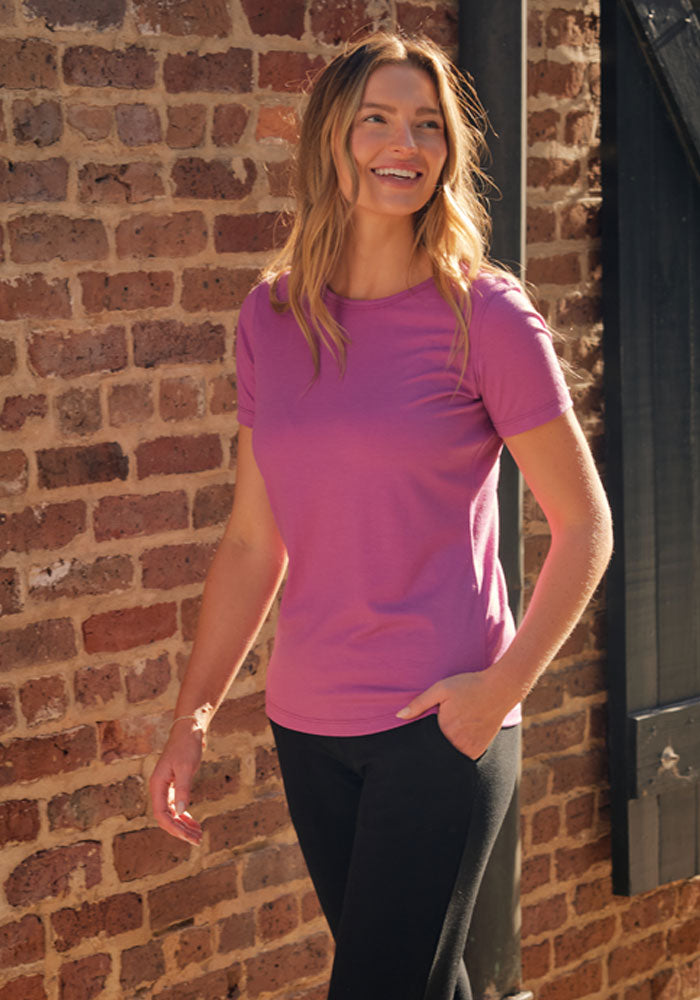 A woman with long, wavy hair smiles as she walks outdoors in sunlight beside a brick wall, wearing the Woolx Liza Crew Neck Tee in Purple Wine and black pants.