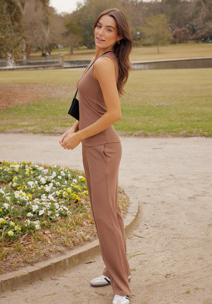 A woman stands on a park path wearing Woolx's Luca Ribbed Pants in Sienna Kiss, paired with a sleeveless brown top and white sneakers. She carries a small black purse and smiles, with flower beds and grass behind her.