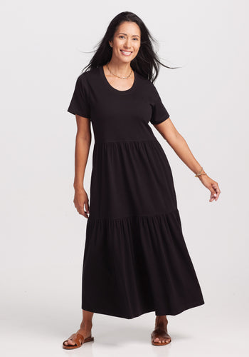 A woman with long black hair smiles in a black short-sleeve, tiered Lucia Dress by Woolx, crafted from temperature-regulating merino wool. She's on a white background, accessorized with a necklace and bracelet, wearing brown sandals. | Denia is 5'8", wearing a size S