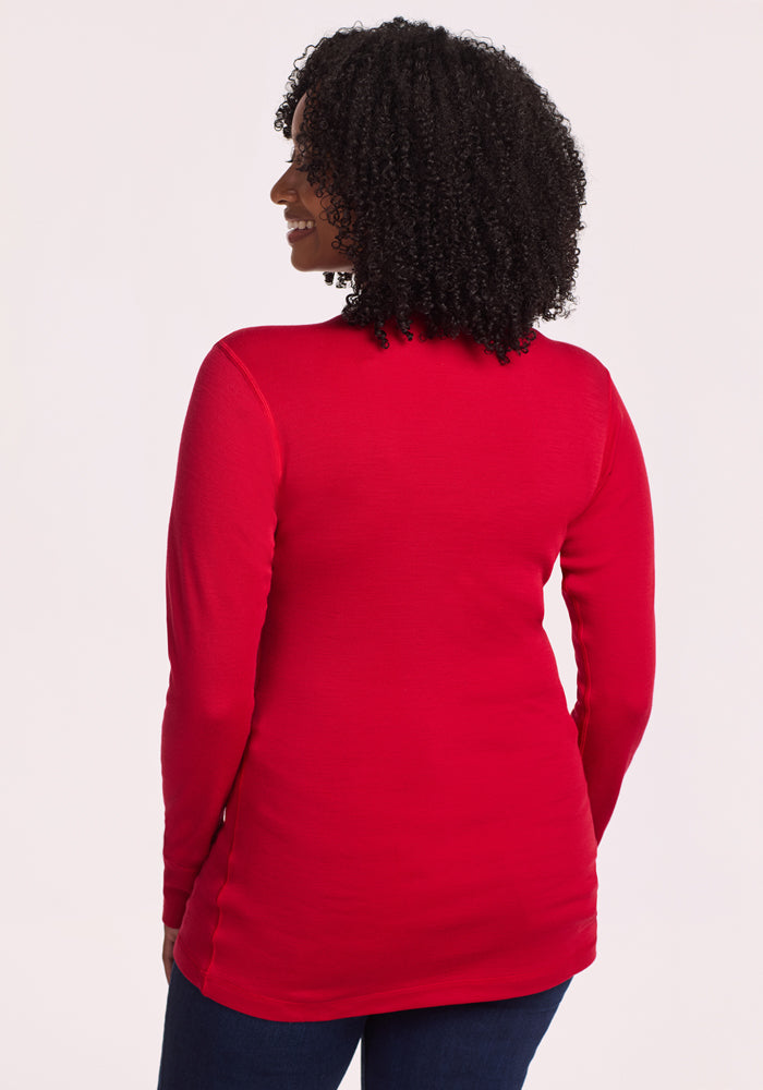 A woman with curly hair faces away from the camera, wearing the Woolx Hannah Long Sleeve Top in Maraschino Cherry and dark jeans against a plain light background.
