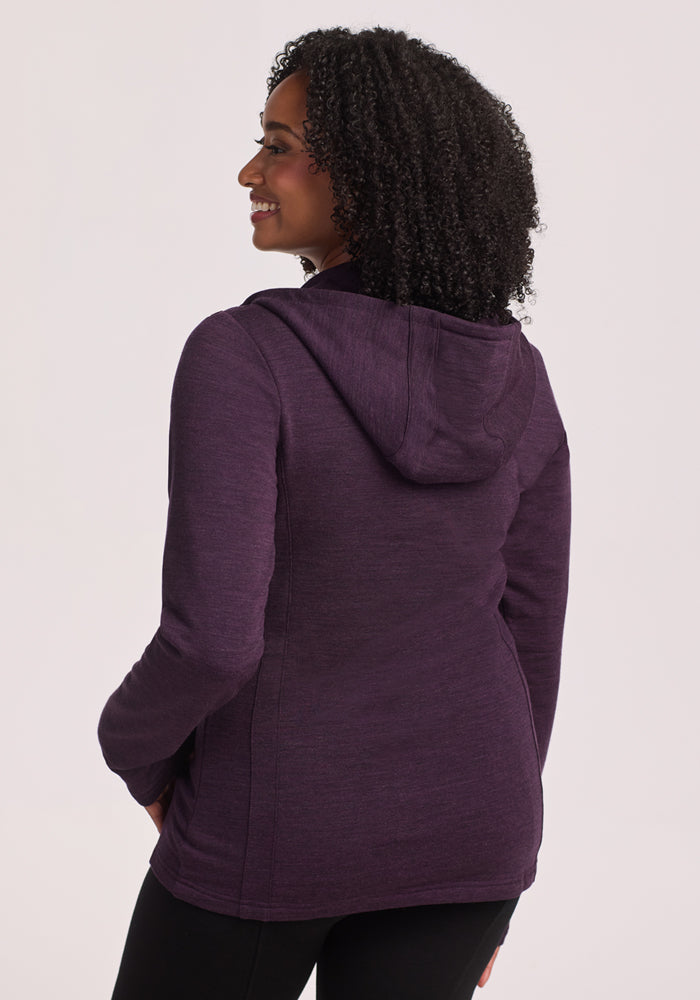 A person with curly hair is smiling and looking to the side, wearing a Woolx Zoey Zip Up Hoodie in Deep Plum and black pants. The photo is taken from behind with a light-colored background.
