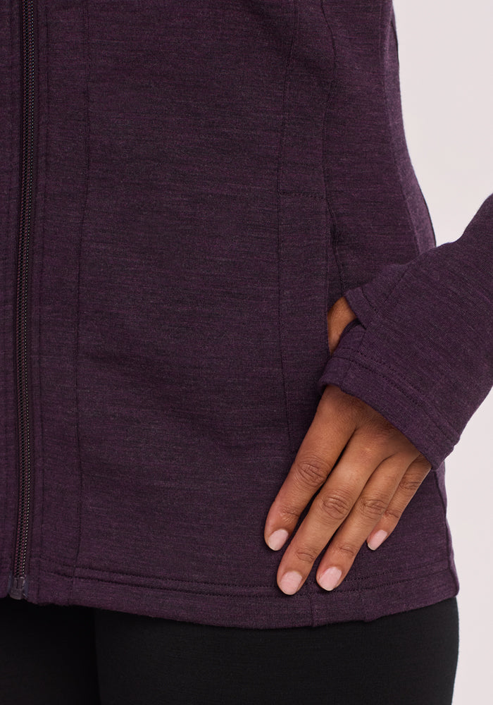 A close-up shows a hand near the side pocket of a Woolx Zoey Zip Up Hoodie in Deep Plum, with the thumb through a sleeve thumb hole and a light, plain background.