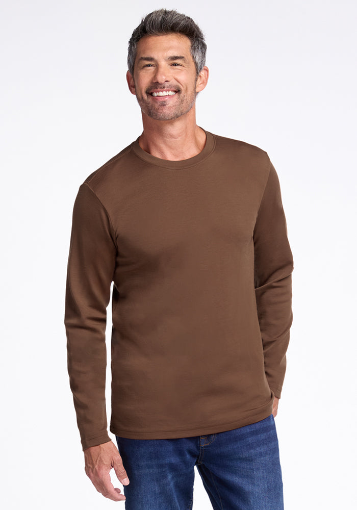 A man with short gray hair and a beard is smiling, wearing the Woolx Easton Crew Neck Shirt in Bourbon Oak and blue jeans, standing against a plain white background. 
