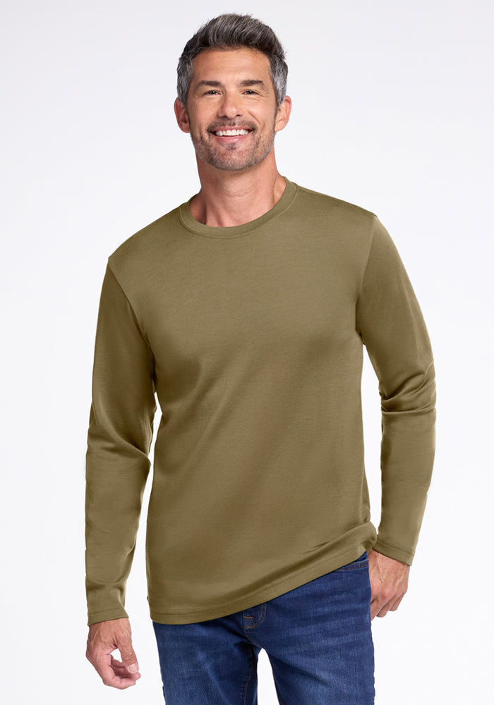 A smiling man with short gray hair wears the Woolx Easton Crew Neck Shirt in Winter Moss and blue jeans, standing against a plain white background. 
