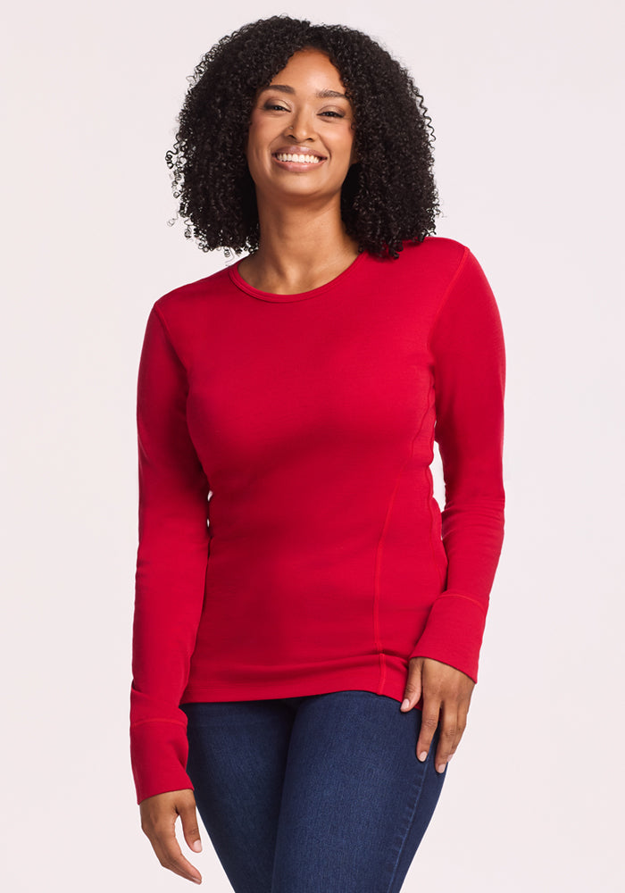 A woman with curly hair smiles in the Woolx Hannah Long Sleeve Top in Maraschino Cherry and dark blue jeans, standing against a plain light background. The merino wool keeps her comfortable with its temperature-regulating fabric. 