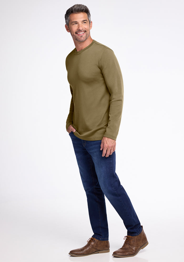 A man with short dark hair and a beard smiles with one hand in his pocket, wearing the Woolx Easton Crew Neck Shirt in Winter Moss, dark blue jeans, and brown boots against a plain white background.
