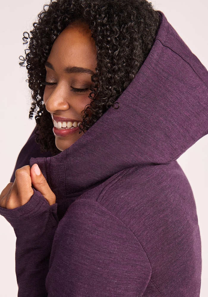 A smiling woman with curly hair wears the Woolx Zoey Zip Up Hoodie in Deep Plum, holding the hood close to her face and looking cozy and content in the soft comfort of Australian Merino wool.