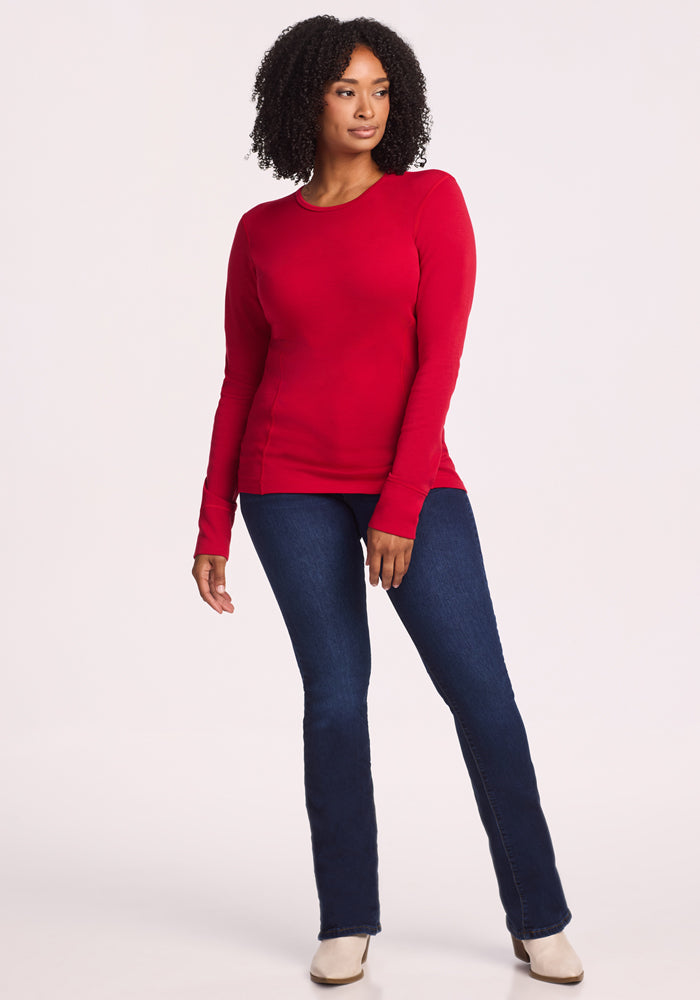 A woman with curly hair wears the Woolx Hannah Long Sleeve Top in Maraschino Cherry, paired with dark blue jeans and beige heeled shoes, standing against a plain light background in a relaxed pose.