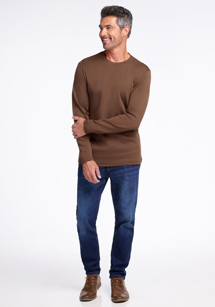 A man wearing a Woolx Easton Crew Neck Shirt in Bourbon Oak, blue jeans, and brown shoes stands smiling against a plain white background.