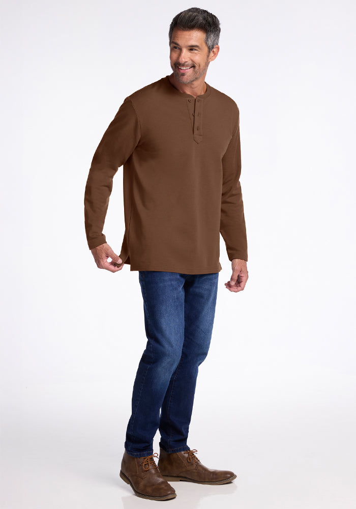 A man with short gray hair smiles in the Woolx Jackson Henley - Bourbon Oak. He pairs the long-sleeve brown merino wool shirt with blue jeans and brown shoes, standing against a plain white background.