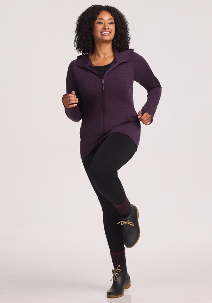A woman with curly hair smiles while lifting one knee, wearing the Woolx Zoey Zip Up Hoodie in Deep Plum, black leggings, dark socks, and black lace-up boots against a light background.