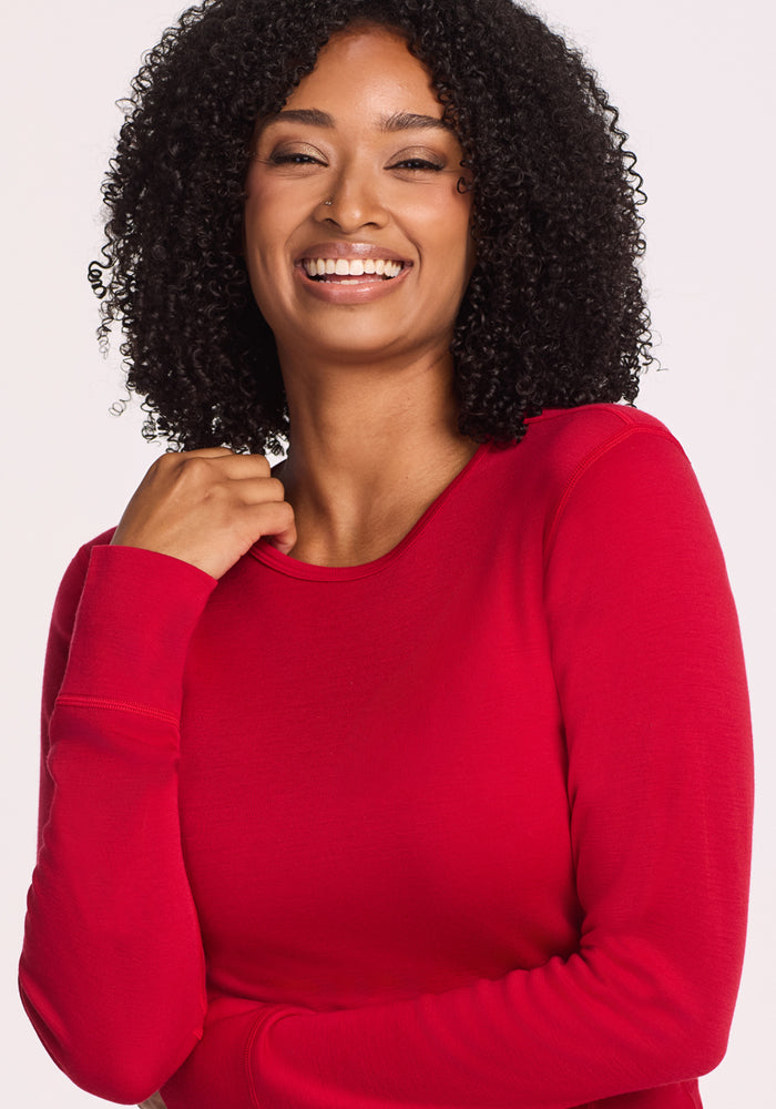 A woman with curly black hair smiles warmly at the camera, wearing the Woolx Hannah Long Sleeve Top in Maraschino Cherry, a temperature-regulating merino wool shirt, against a plain light background.