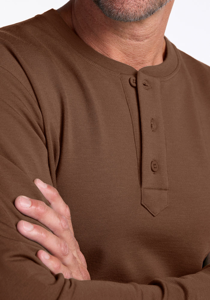 A close-up of a man wearing the Woolx Jackson Henley - Bourbon Oak, a brown merino shirt with three buttons, standing with his arms crossed. Only the lower part of his face is visible.