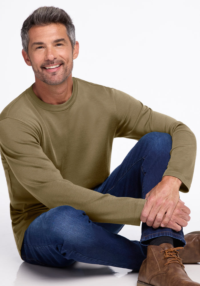 A smiling man with short gray hair and a trimmed beard sits casually on the floor wearing Woolx’s Easton Crew Neck Shirt in Winter Moss, blue jeans, and brown lace-up boots against a plain white background.