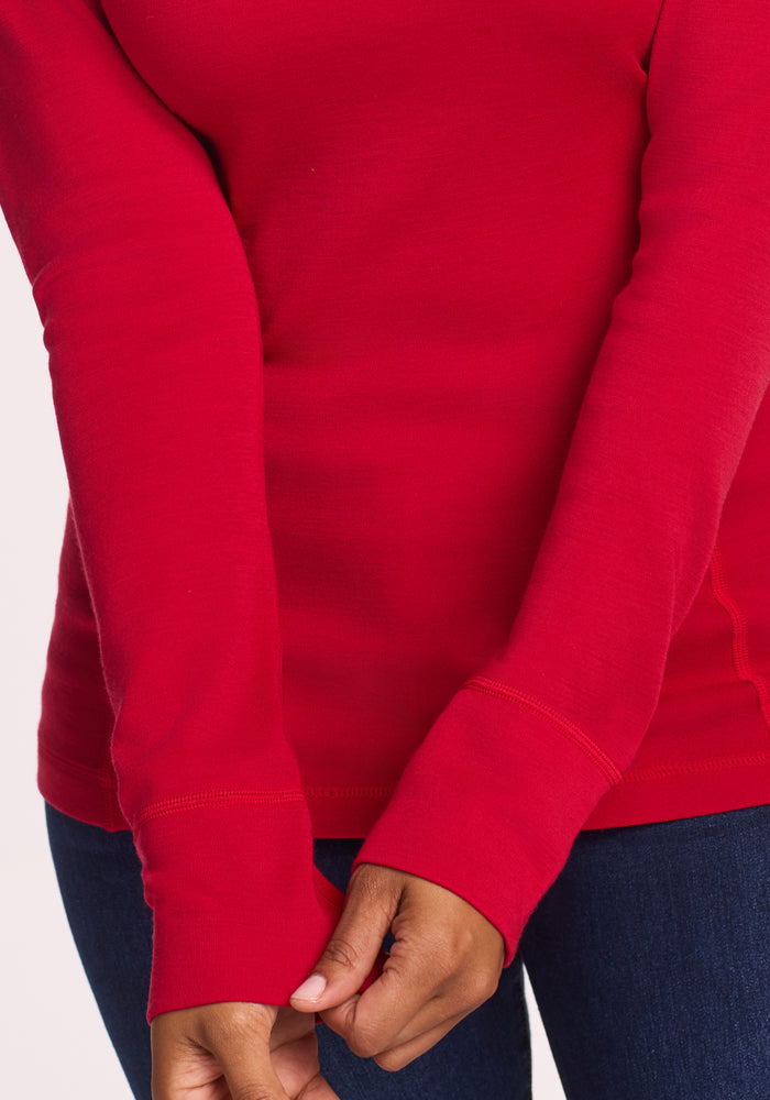 A person wearing the Woolx Hannah Long Sleeve Top in Maraschino Cherry with dark blue jeans, standing with arms crossed and holding one sleeve cuff. Only their torso and arms are shown.
