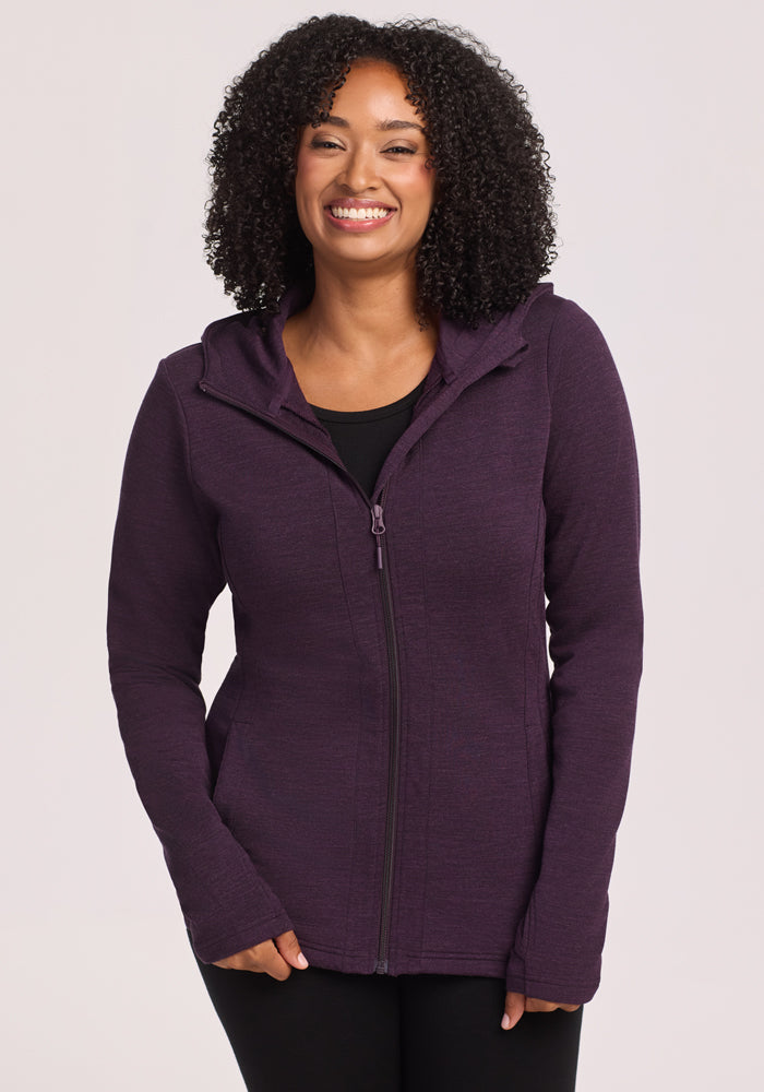 A woman with curly hair smiles while wearing a Woolx Zoey Zip Up Hoodie in Deep Plum over a black top, standing against a plain light background. 