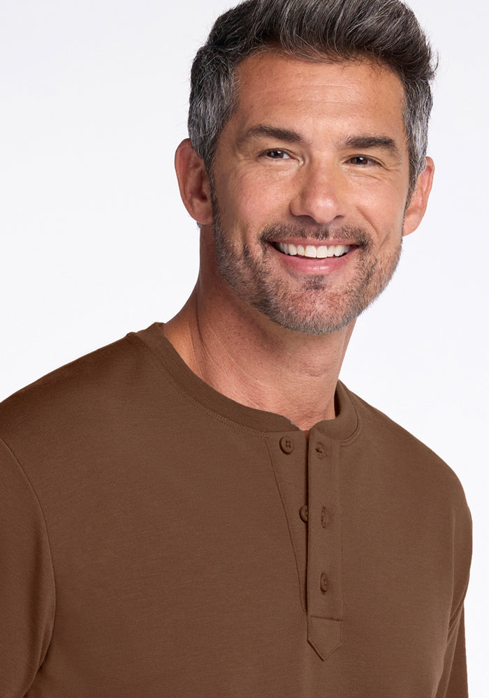 A middle-aged man with short, graying hair and a trimmed beard smiles while wearing the Woolx Jackson Henley in Bourbon Oak against a plain, light background.