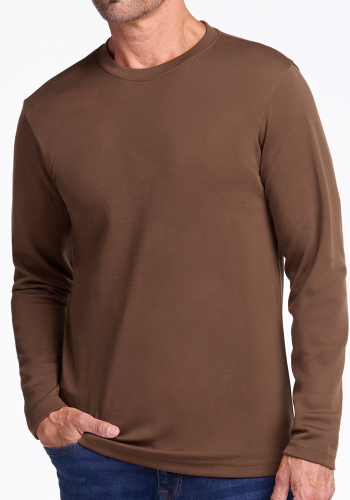 A man wearing the Woolx Easton Crew Neck Shirt in Bourbon Oak and blue jeans stands with one hand in his pocket against a white background.