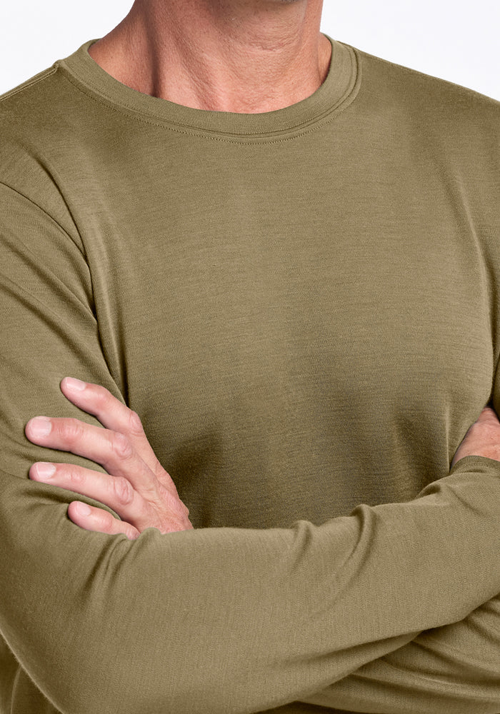 A person wears the Woolx Easton Crew Neck Shirt in Winter Moss, an olive green long sleeve merino top, standing with crossed arms against a plain white background.