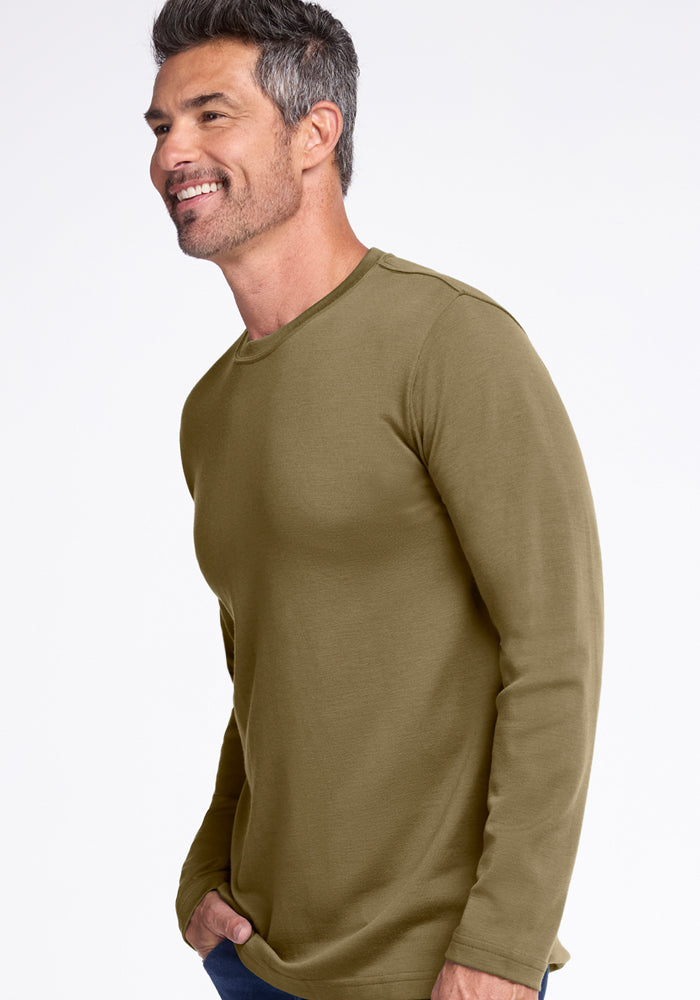 A smiling man with short salt-and-pepper hair wears the Woolx Easton Crew Neck Shirt in Winter Moss and jeans, standing against a plain white background and looking to his left.