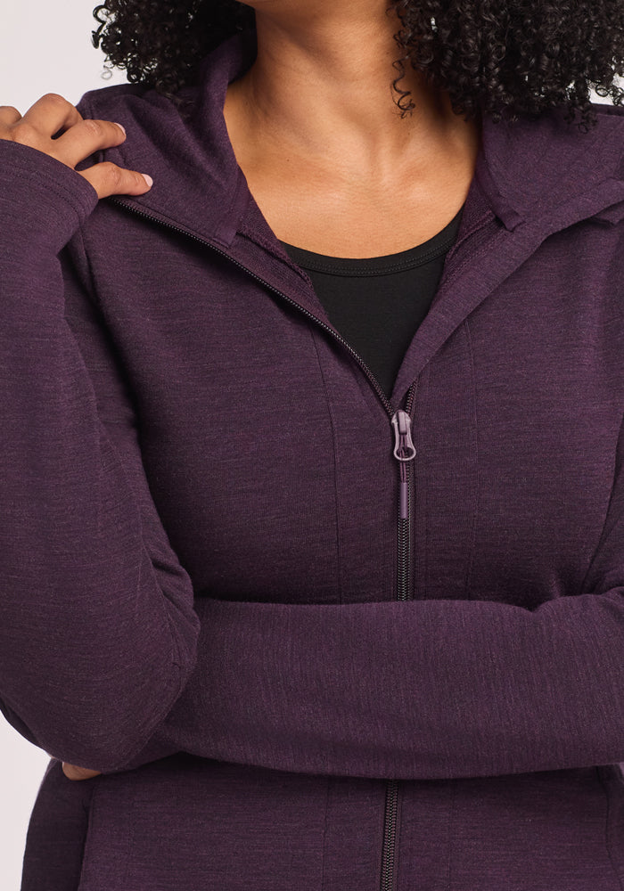 A woman with curly hair wears the Woolx Zoey Zip Up Hoodie in Deep Plum over a black top, touching the collar with her left hand. Her face is partially out of frame.