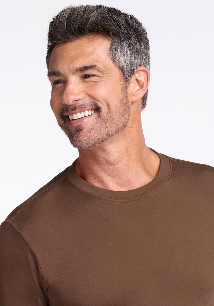 A man with short salt-and-pepper hair and a trimmed beard smiles to the side, wearing the Woolx Easton Crew Neck Shirt in Bourbon Oak against a light background.