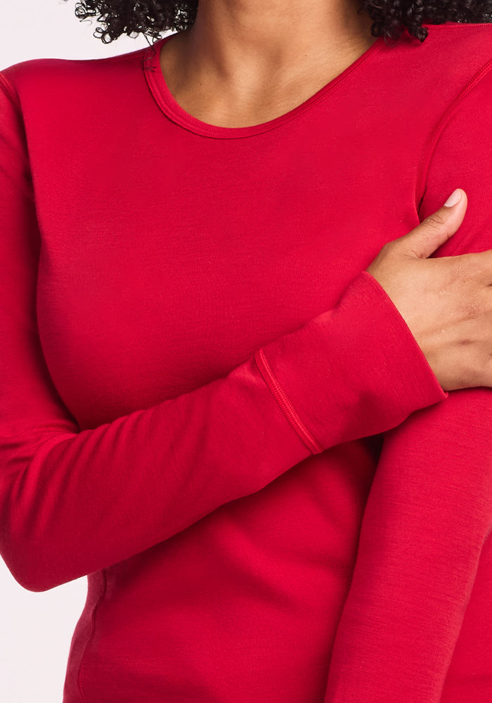 A person with curly hair wears the Woolx Hannah Long Sleeve Top in Maraschino Cherry, folding one arm across their body. The torso-focused image highlights the shirt’s temperature-regulating comfort.