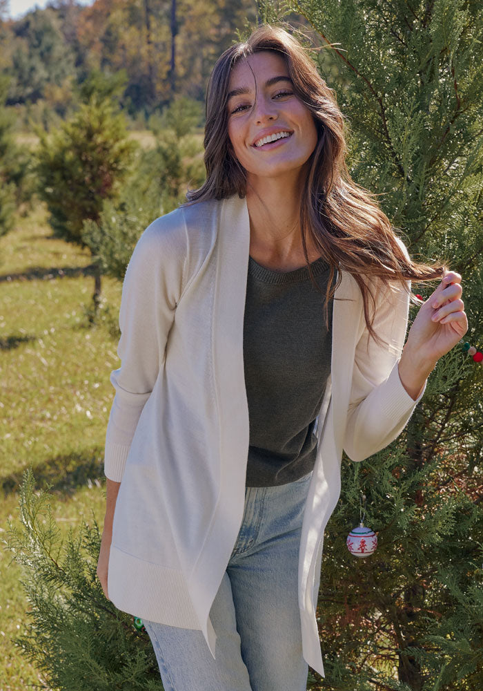 A woman with long brown hair smiles outdoors beside a decorated evergreen tree, wearing the Woolx Macie Sweater in Dewdrop Pearl over a dark top and jeans, with a sunny green landscape in the background.