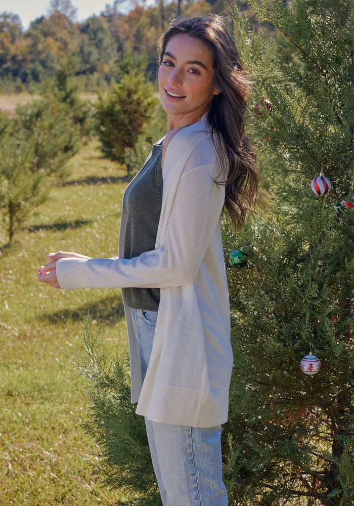 A woman with long brown hair smiles outdoors near a decorated evergreen, wearing the Woolx Macie Sweater in Black over a dark top and jeans. Green grass and trees with a clear sky complete the background.