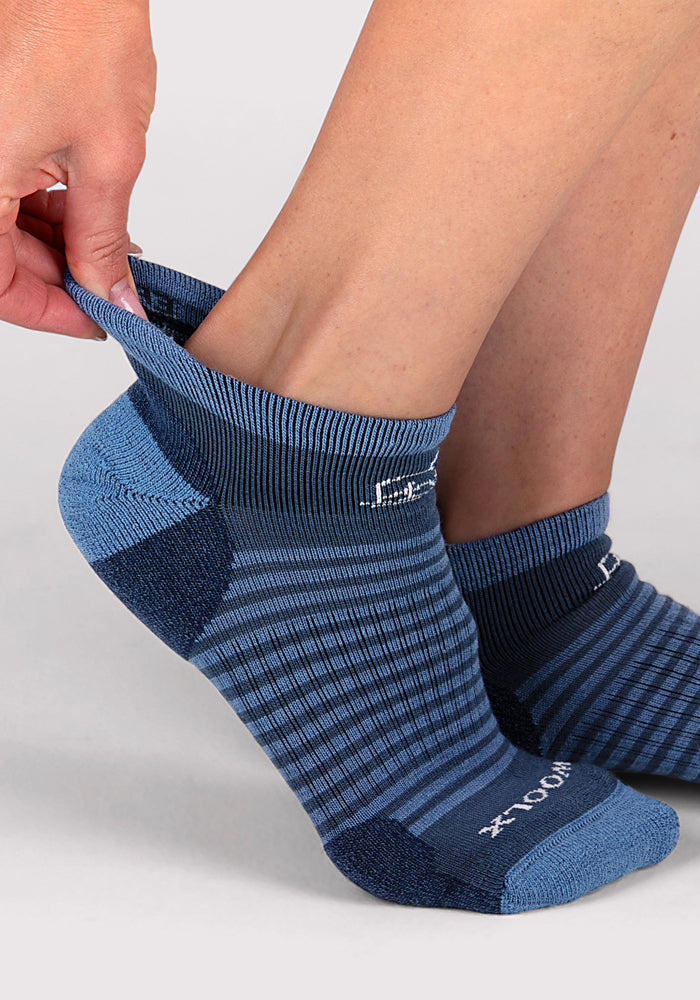 A person pulls on a Woolx Womens Amelia No Show Sock in Majolica Blue Stripe, with the matching sock already worn, set against a neutral background.
