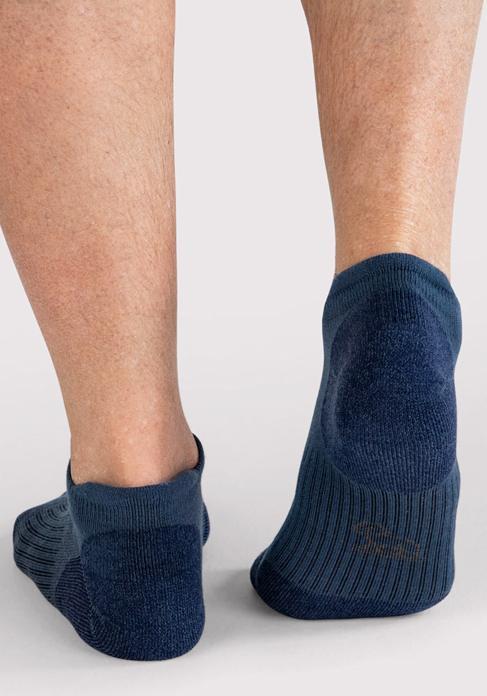 A close-up of the back of a person’s lower legs and feet wearing Woolx Mens Rowan No Show Socks in Majolica Blue. The plain, light-colored background highlights the dark blue, moisture-wicking socks.