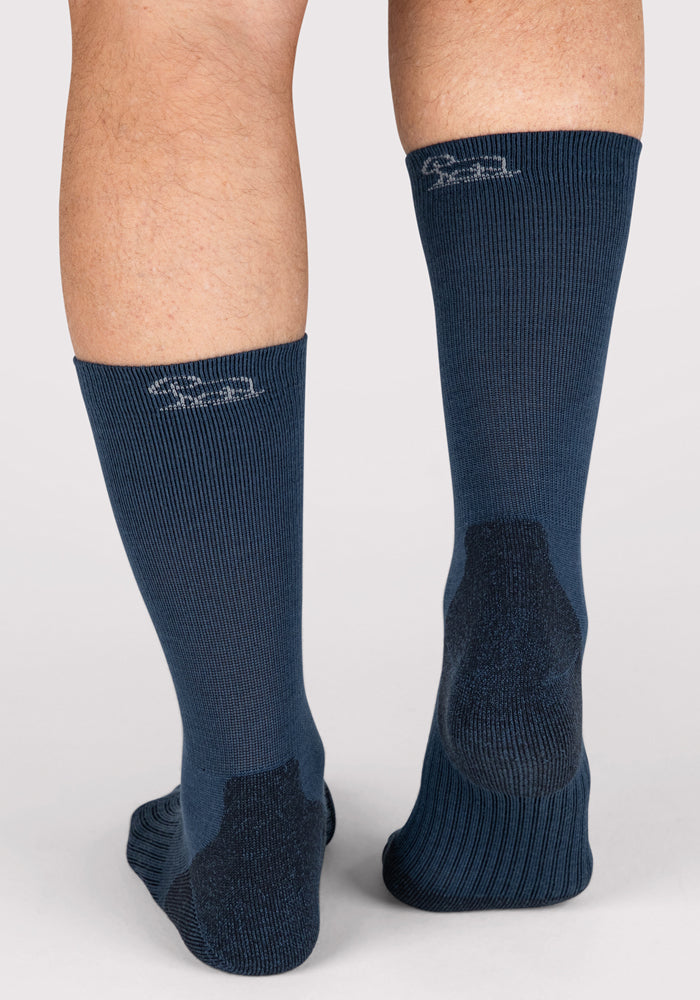 A close-up of two feet from behind, wearing Woolx Mens Harbor Crew Socks in Majolica Blue. The ribbed Merino wool socks feature a subtle logo near the top back of each calf against a plain, light background.