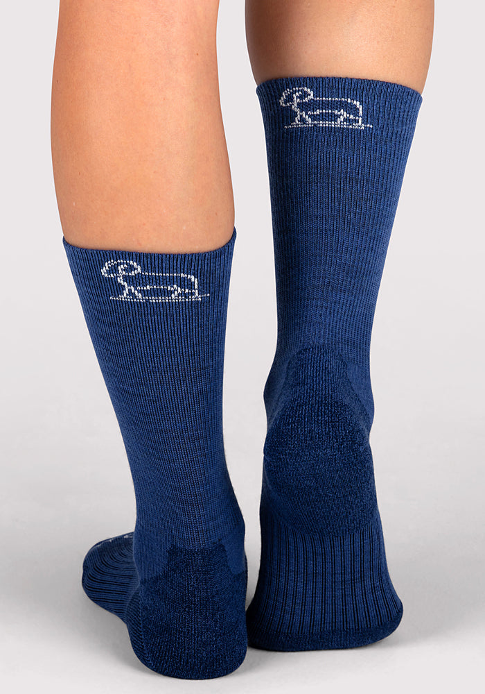 A close-up of the back of a person's lower legs wearing Woolx Womens Harbor Crew Sock in Sailor Blue, featuring a small white dachshund design near the top. The background is plain and light-colored.