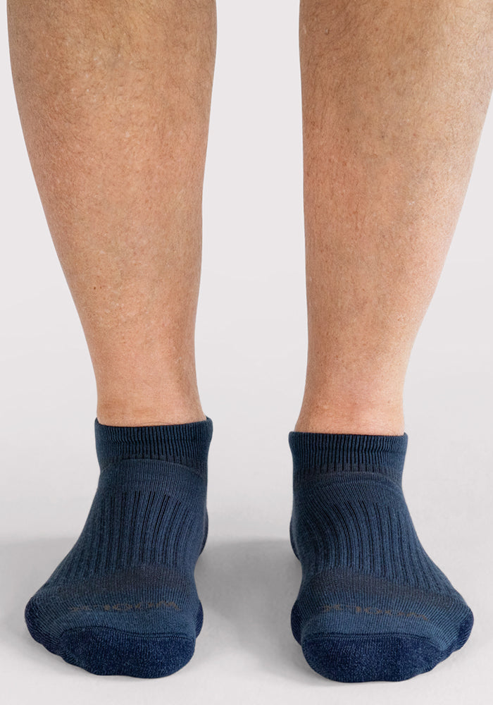 A close-up of a person’s lower legs and feet wearing Woolx Mens Rowan No Show Socks in Majolica Blue, standing on a light surface with a plain background.