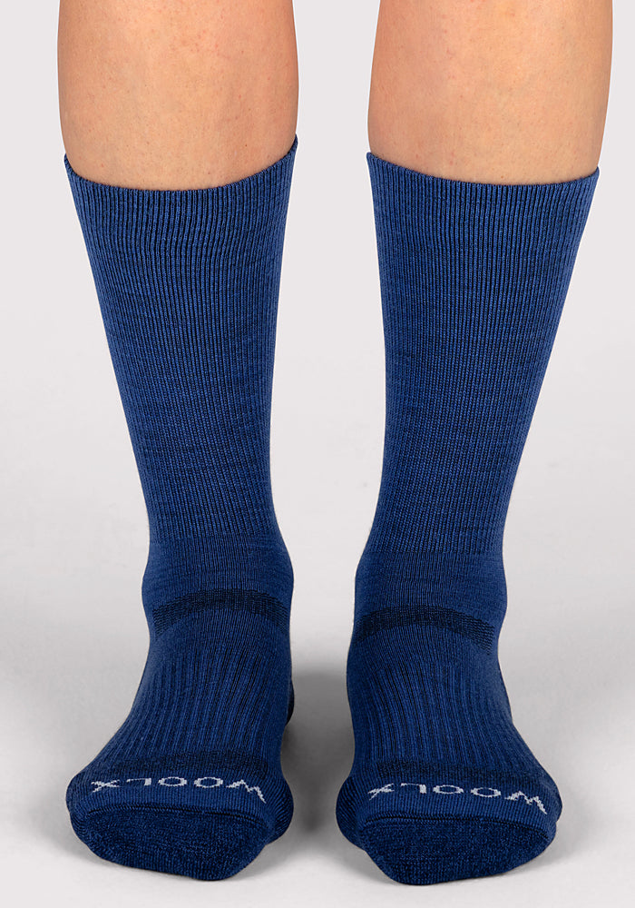 A pair of feet in Woolx Womens Harbor Crew Socks in Sailor Blue, with a ribbed texture and "WOOLX" in white near the toes, stand on a light gray surface. These Merino wool socks provide natural odor-resistant comfort.