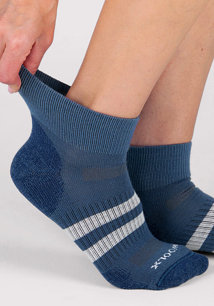 A woman wearing Woolx Womens Rambler Ankle Socks in Majolica Blue with white stripes adjusts one sock higher on her foot. The background is plain and light-colored.