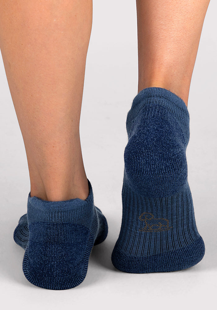Close-up of two feet wearing Woolx Womens Rowan No Show Socks in Majolica Blue, standing on a light gray floor. The left sock is slightly wrinkled at the heel, and the right features a faint design near the sole.