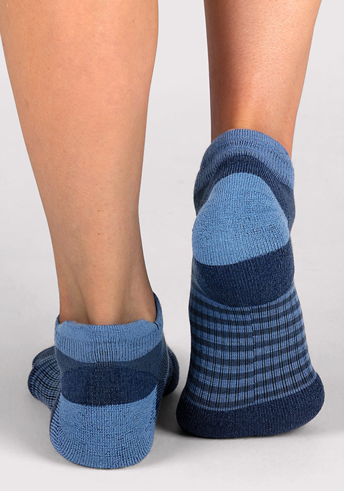 A close-up of two feet wearing Woolx Womens Amelia No Show Socks in Majolica Blue Stripe, standing on a light gray surface.