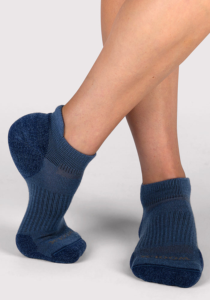 A woman wearing Woolx Womens Rowan No Show Socks in Majolica Blue stands with one foot crossed in front of the other against a light grey background.