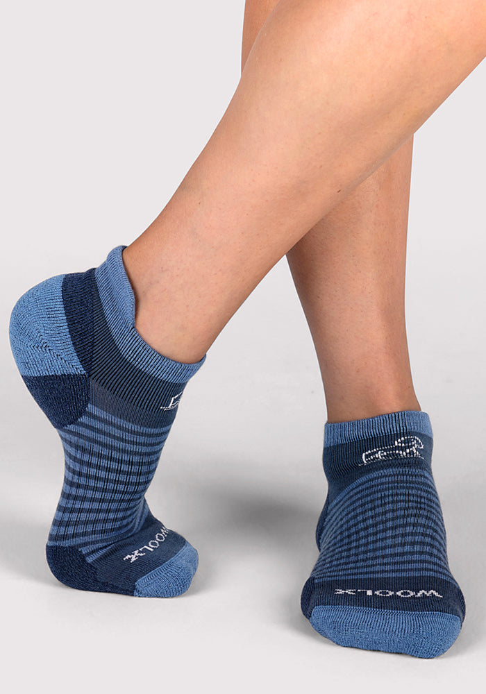 A person stands on tiptoe against a light grey background, showcasing the stretch and fit of Woolx Womens Amelia No Show Socks in Majolica Blue Stripe.