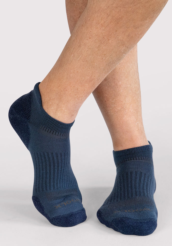 A close-up shows a person’s feet crossed, wearing Woolx Mens Rowan No Show Socks in Majolica Blue. Made from Merino wool, these cushioned socks deliver all-day comfort and moisture-wicking performance against a plain background.