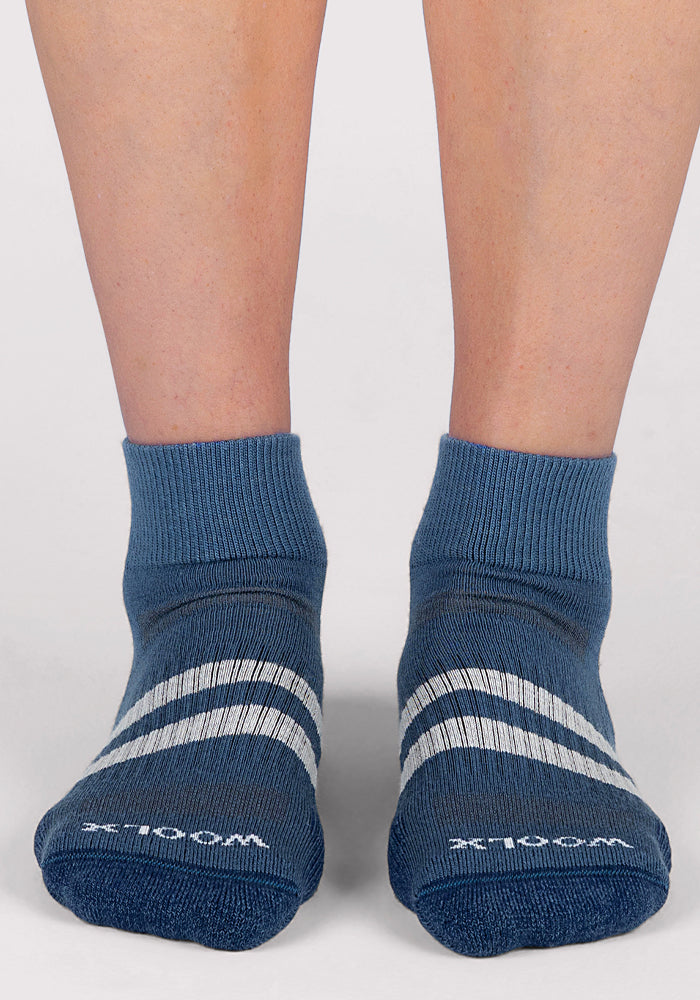 A person wearing Woolx Womens Rambler Ankle Sock in Majolica Blue with white diagonal stripes stands on a light-colored surface. The moisture-wicking socks have white text on the toes, and the legs are visible up to mid-calf.