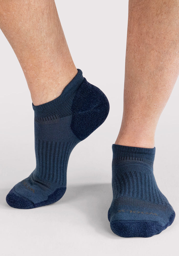 Close-up of a person wearing Woolx Mens Rowan No Show Socks in Majolica Blue. Featuring ribbed, moisture-wicking fabric and reinforced toes and heels, one foot is slightly raised on a light surface.