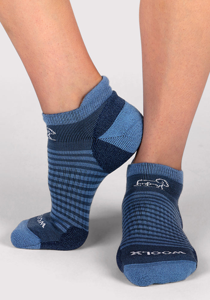 Close-up of a person wearing Woolx Womens Amelia No Show Socks in Majolica Blue Stripe, featuring breathable fabric, horizontal blue stripes, and white logos and text near the toes and ankles.
