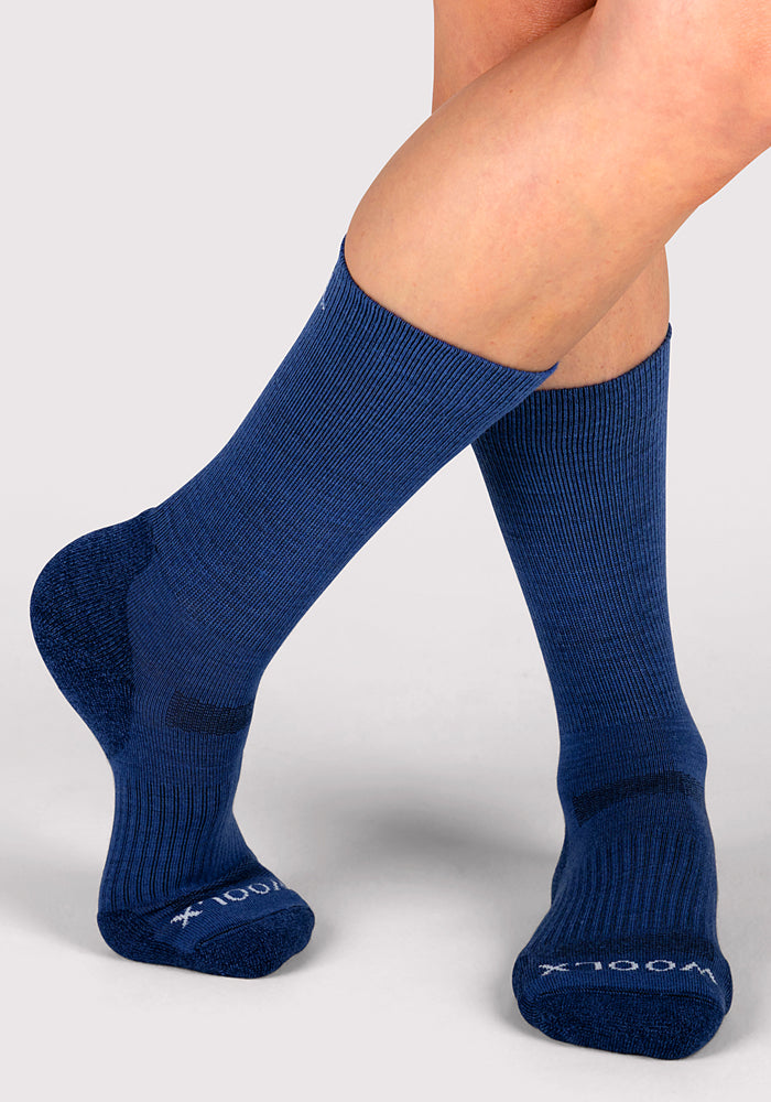 A person wears Woolx Womens Harbor Crew Sock in Sailor Blue, featuring a mid-calf fit, moisture-wicking fabric, ribbed texture, and "x-100" printed in white on the toes, standing on a light gray background.