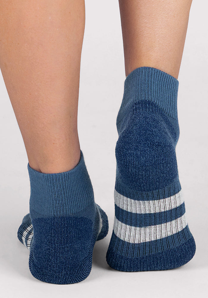 A close-up shows two feet in Woolx Womens Rambler Ankle Sock - Majolica Blue, featuring moisture-wicking fabric and white with dark blue striped soles, standing on a light gray surface.