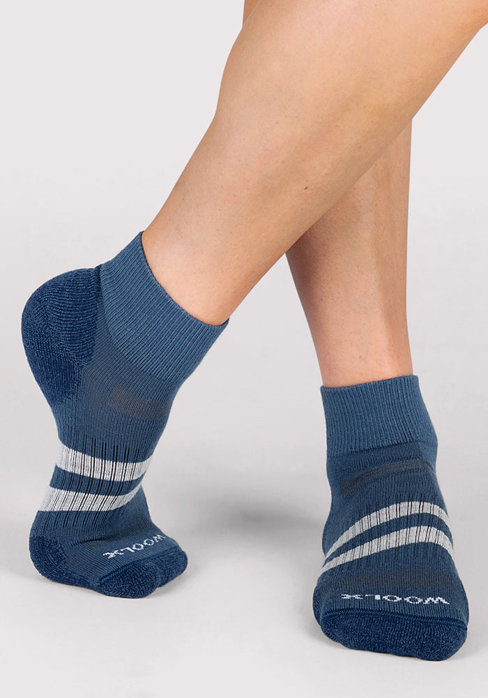 A person stands on one foot, wearing Woolx Womens Rambler Ankle Sock in Majolica Blue. These athletic socks feature white diagonal stripes, a textured pattern, and offer moisture-wicking performance.