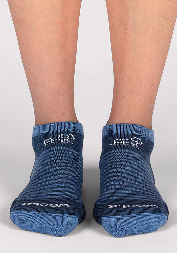 A pair of feet in Womens Amelia No Show Socks by Woolx in Majolica Blue Stripe, featuring "WOOL" on the left and "RICH" on the right, standing on a light gray surface.