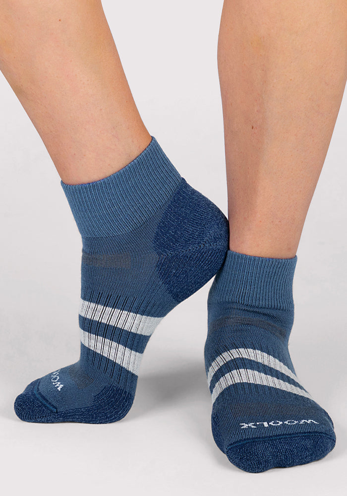 Close-up of a person wearing Woolx Womens Rambler Ankle Sock in Majolica Blue, featuring white stripes, ribbed cuffs, textured heel and toe, and moisture wicking Merino wool. Socks shown on a light gray surface.